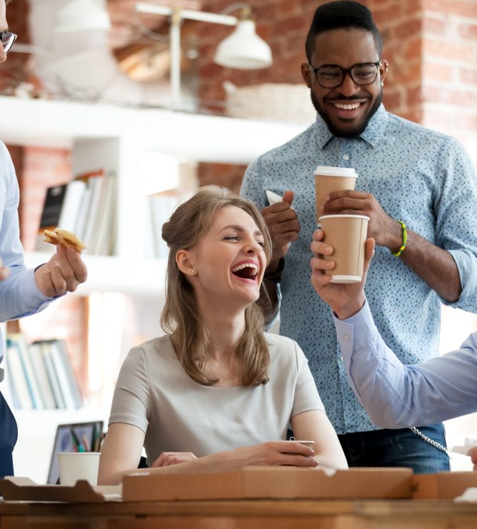 Happy diverse colleagues have fun at lunch break in office, smiling multiracial employees laugh and talk eating pizza and drinking coffee, excited workers celebrate shared win ordering takeaway food