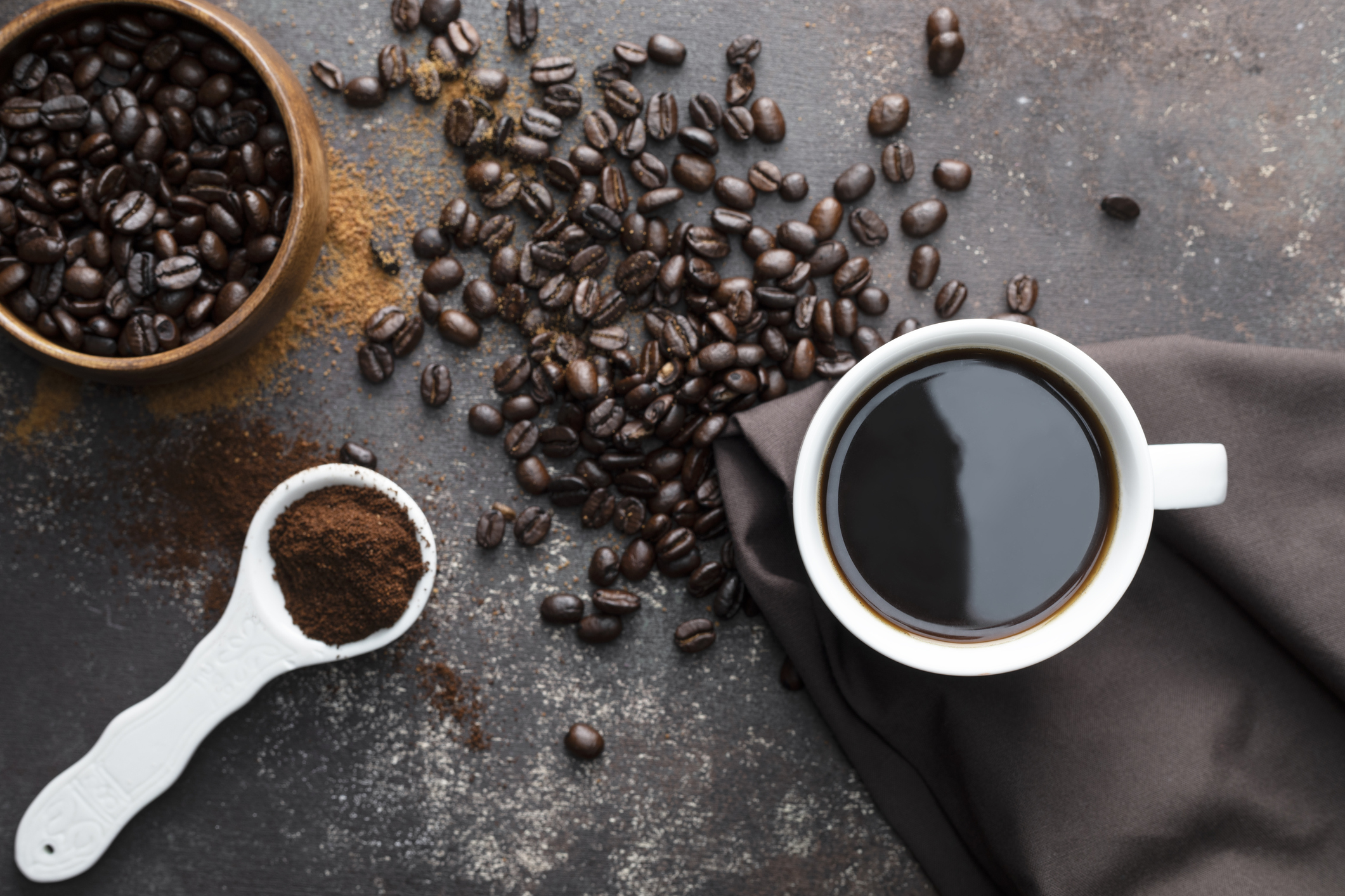 Direct above view of roasted coffee beans, white coffee cup with black coffee, dark brown colored napkin with one white spoon full with blended coffee on dark brown background.