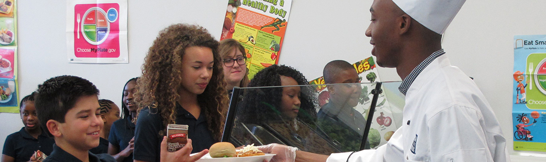 chef hands plate of food to accepting students in cafeteria full of students